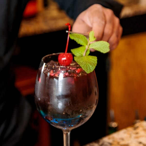 A hand garnishes a dark Indian cocktail with a cherry and mint sprig in a wine glass.