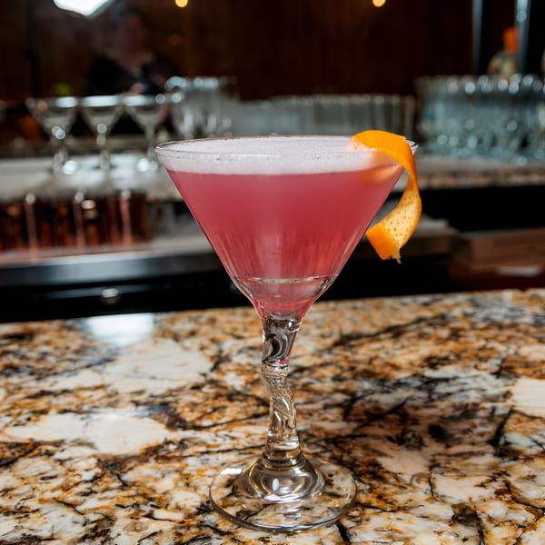 Pink cocktail with foam and an orange peel garnish on a marble bar top.
