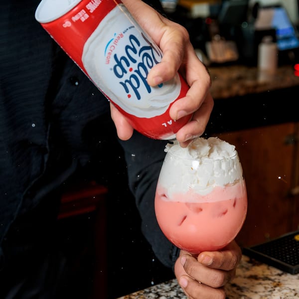 A bartender adds whipped cream from a can to a pink, iced cocktail in a wine glass.