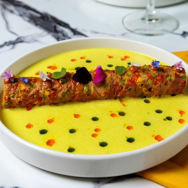 A colorful kebab in a yellow sauce, garnished with edible flowers and herbs, in a white bowl.