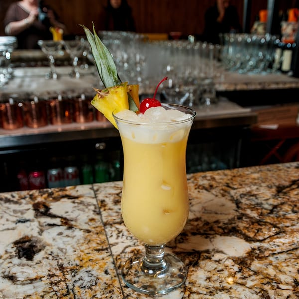 A yellow cocktail with ice, a cherry, and pineapple garnish sits on a marble bar.