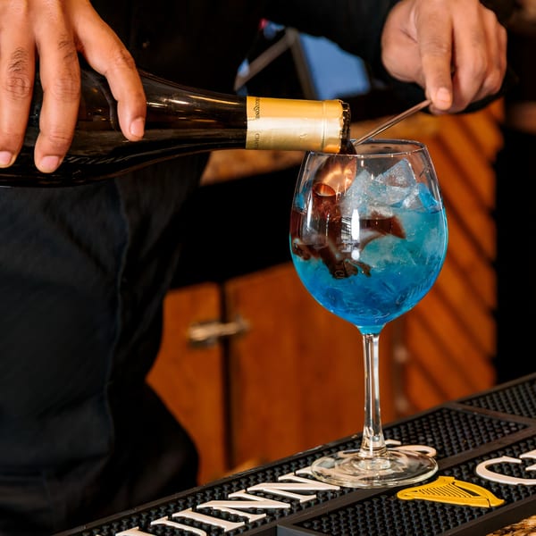 A bartender pours red wine from a bottle into a blue cocktail with ice in a large glass.