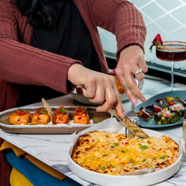 A person cuts into a cheesy naan bread, surrounded by other Indian dishes and a cocktail.