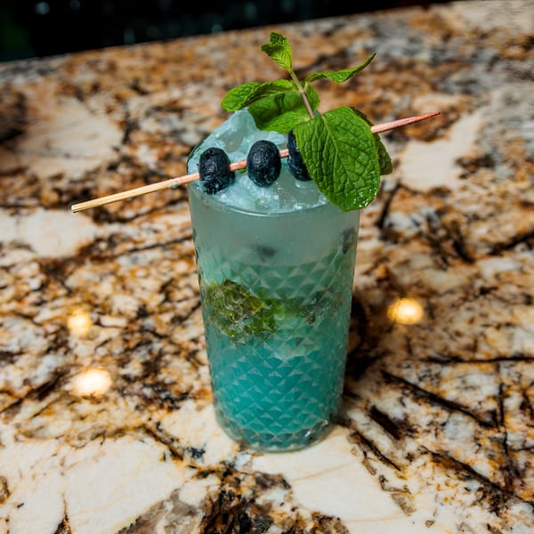Blue cocktail with crushed ice, blueberries on a skewer, and mint sprigs on a marble counter.