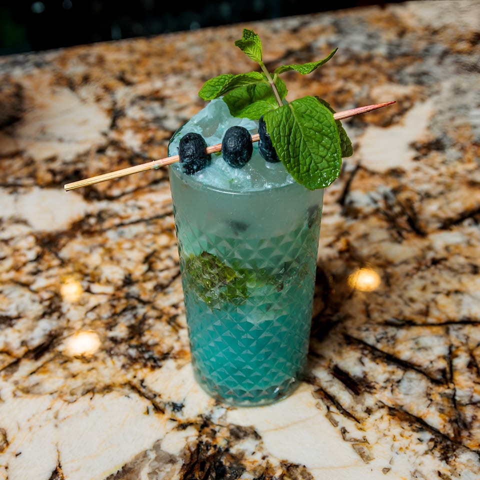 Blue cocktail with crushed ice, blueberries on a skewer, and mint sprigs on a marble counter.