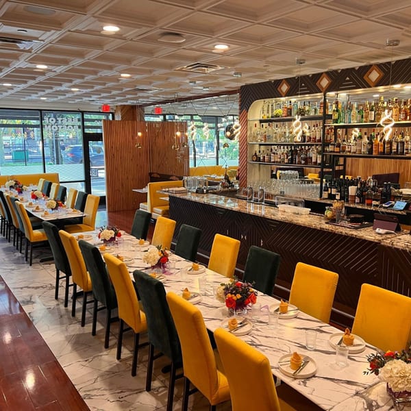 Elegant restaurant interior with long dining tables, yellow and black chairs, and a well-stocked bar.