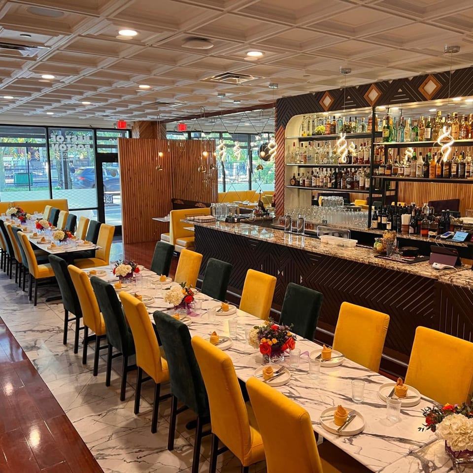 Elegant restaurant interior with long dining tables, yellow and black chairs, and a well-stocked bar.