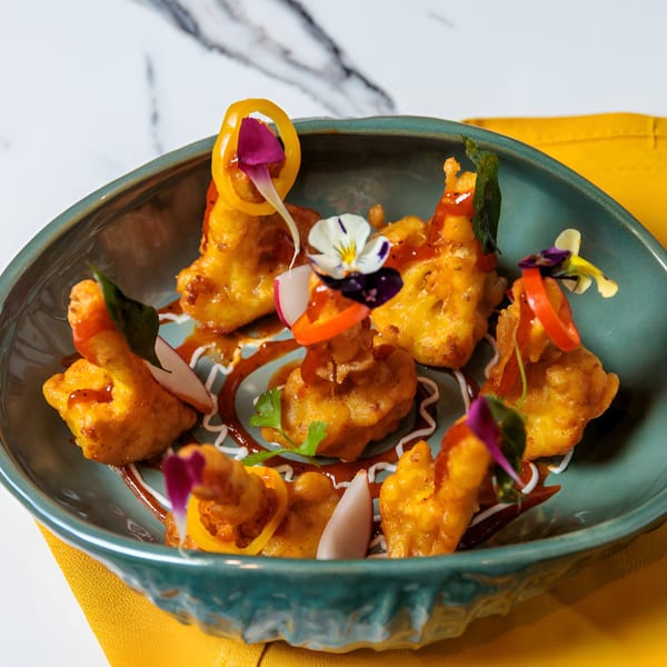 Golden fried cauliflower bites with sauce, edible flowers, and peppers in a teal bowl.