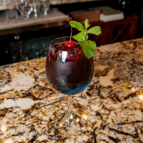 Dark red cocktail in a stemmed glass, garnished with a cherry and mint, on a marble bar.