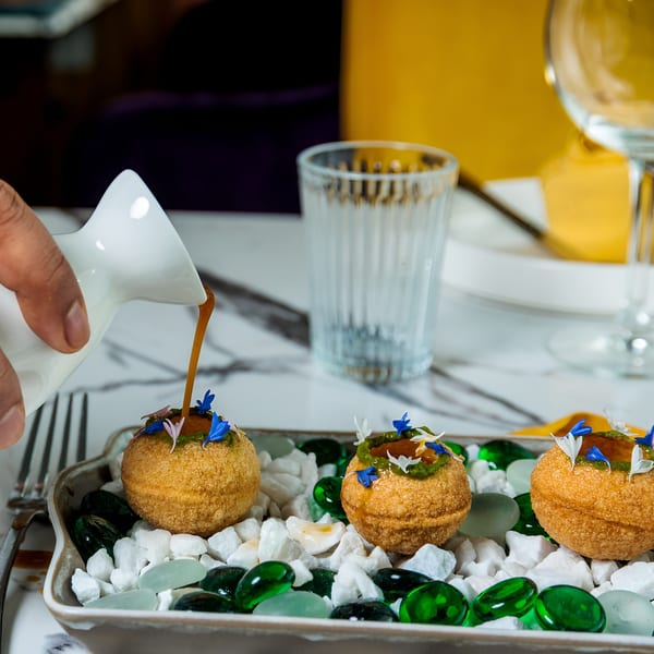 A hand pours sauce into a golden-fried dish garnished with blue and white petals, served on decorative stones.