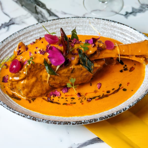 Lamb shank in a rich orange sauce, garnished with pink petals, herbs, and red chilies.