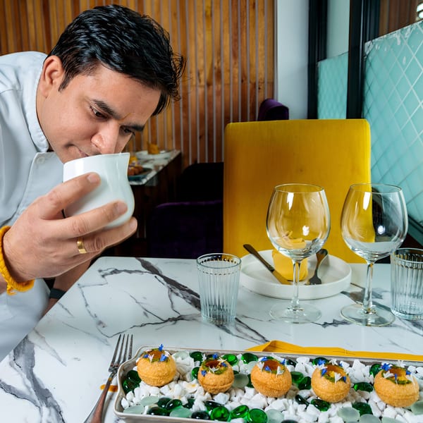 A chef in a white uniform smelling a white pitcher, with appetizers and wine glasses on a marble table.