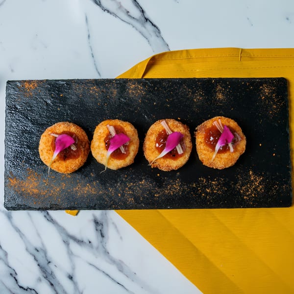 Four golden-brown appetizers with red jam and pink petals on a black slate, on a marble table with a yellow napkin.