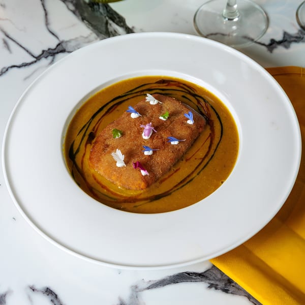 A rectangular fried dish with edible flowers in a bowl of orange soup with dark swirls.