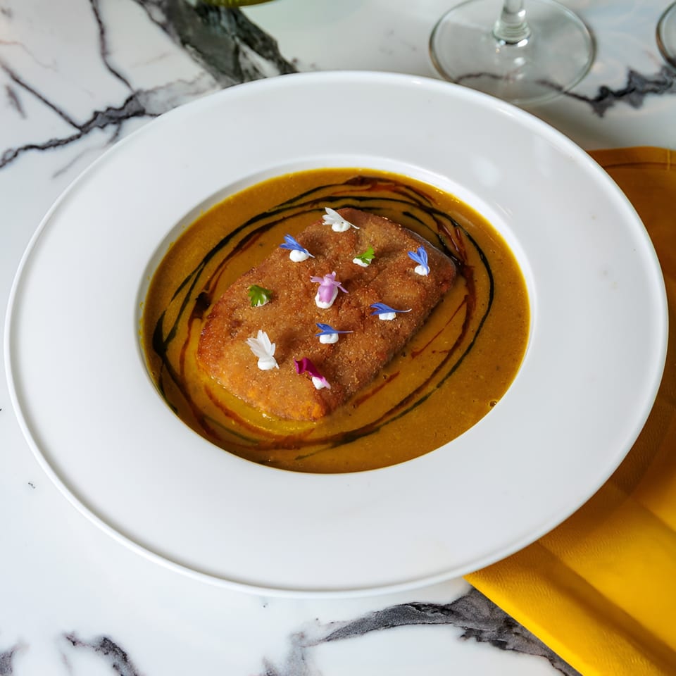 A rectangular fried dish with edible flowers in a bowl of orange soup with dark swirls.