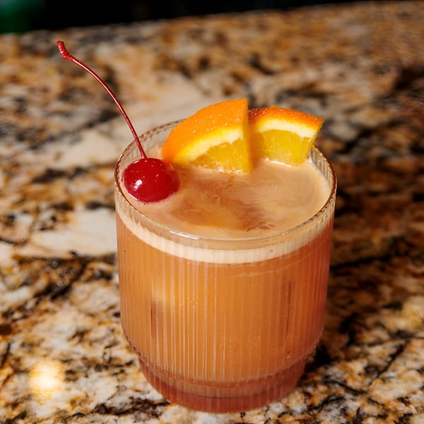 A vibrant orange cocktail with foam, garnished with two orange slices and a red cherry on a marble counter.