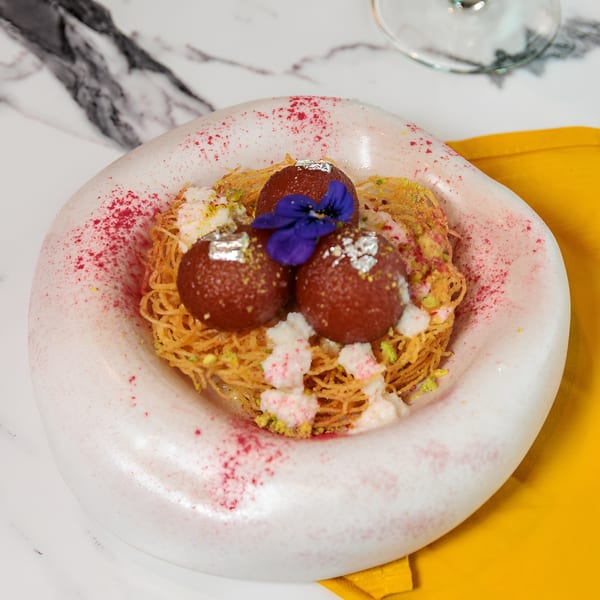 Gulab Jamun dessert with edible silver leaf, purple flower, and vermicelli on a white plate.