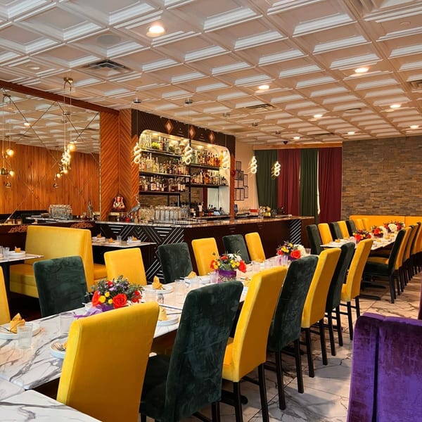 Restaurant interior with long dining tables, yellow and dark green chairs, and a full bar in the background.